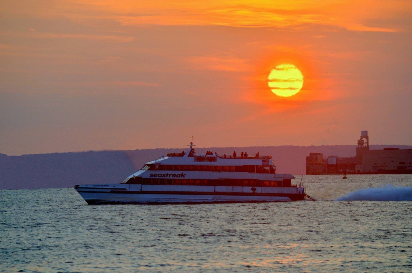A Seastreak ferry cruises through the harbor with a perfect sunset on the horizon.