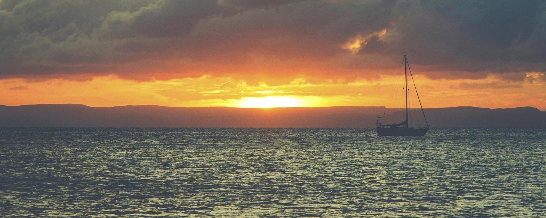 A gorgeous sunset silhouettes a solitary sailboat on the water at dusk.