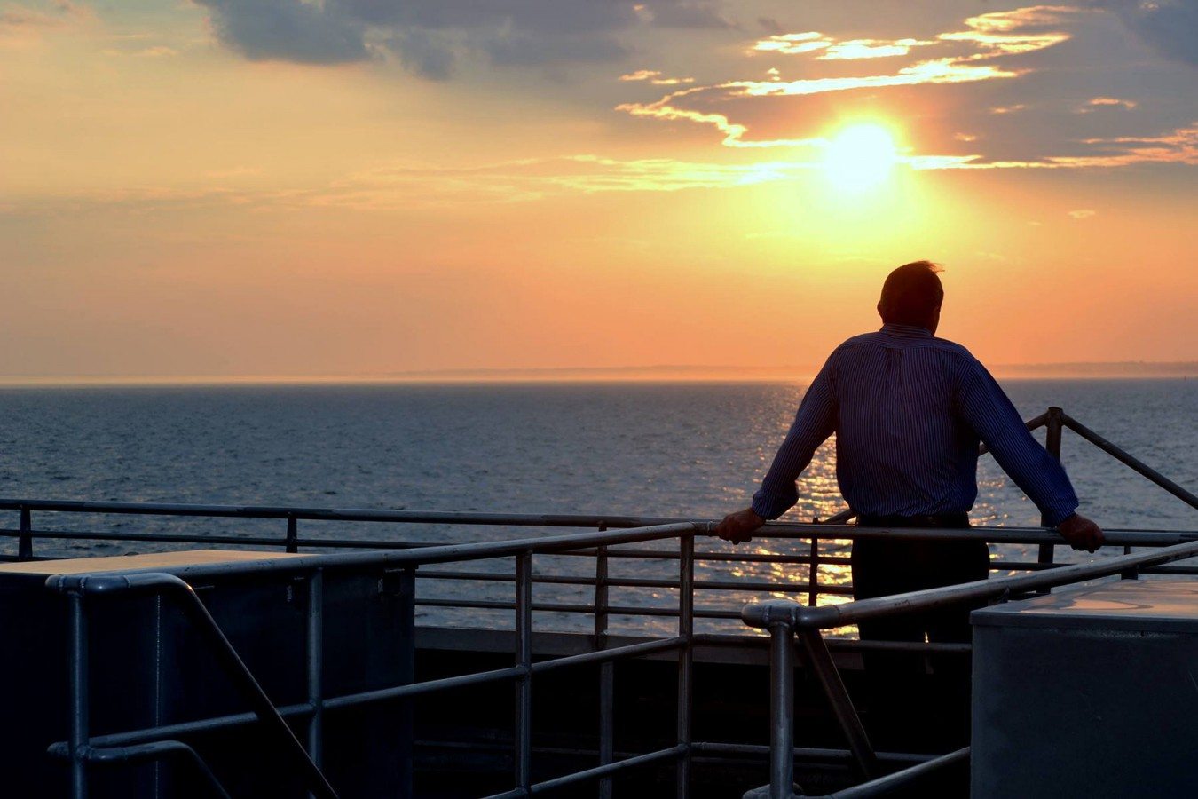 A man looks out over the water from the deck of a Seastreak ferry while the sun sets on the horizon.